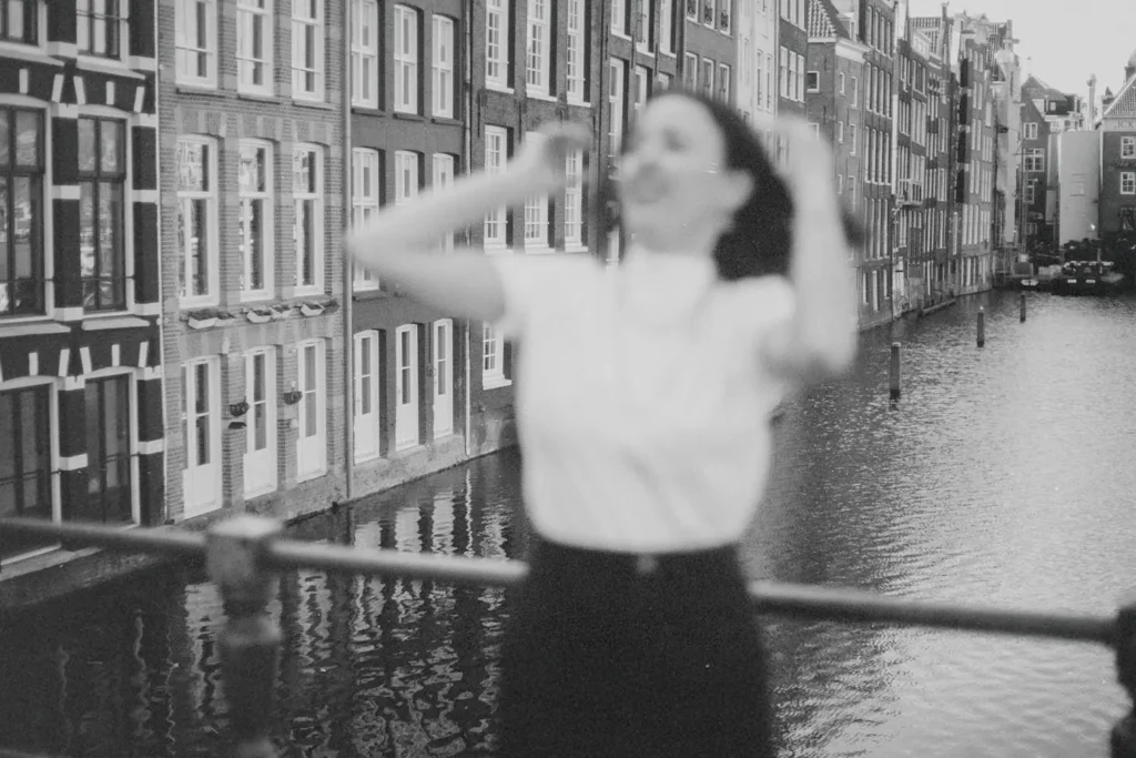 Woman posing on a railing above a waterfront canal, with rows of traditional European buildings in the background.