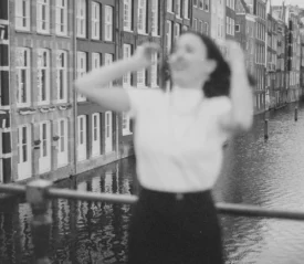 Woman posing on a railing above a waterfront canal, with rows of traditional European buildings in the background.