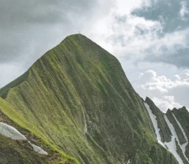 Steep green mountain peak with a small cross at the summit beneath a dramatic cloudy sky