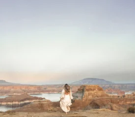 Person in a light-colored dress standing on a cliff edge overlooking a vast canyon landscape at sunrise