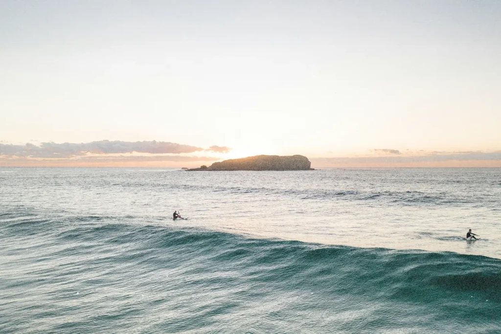 Two surfers sitting on their boards in gentle ocean waves at sunrise with a small island in the distance