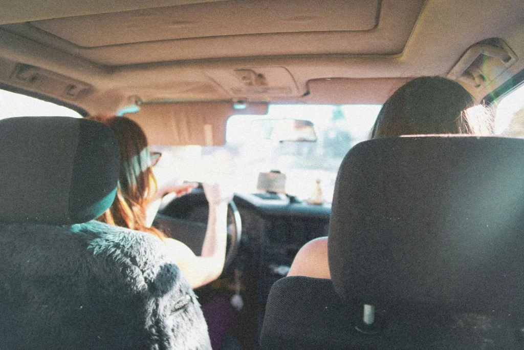 Two people sit in the front seats of a car during daylight, one driving as sunlight streams through the windshield
