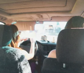 Two people sit in the front seats of a car during daylight, one driving as sunlight streams through the windshield