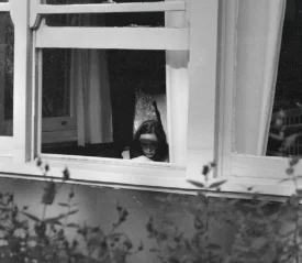 View through an open window into a dimly lit room where a person is seated near a chair, framed by white curtains and surrounding foliage