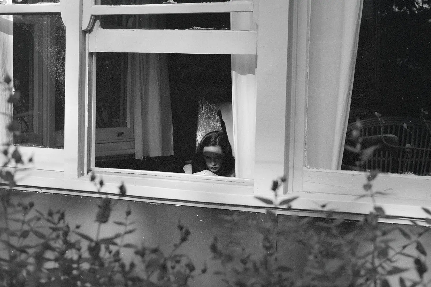 View through an open window into a dimly lit room where a person is seated near a chair, framed by white curtains and surrounding foliage