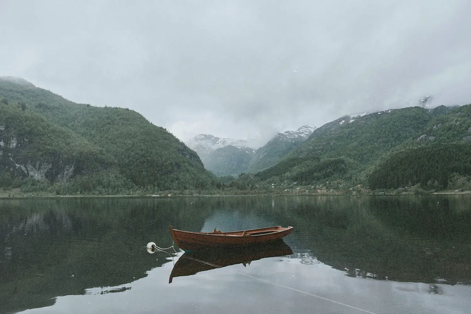 Wooden rowboat floating on a still lake surrounded by steep, forested mountains beneath an overcast sky