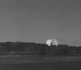 Large satellite dishes rising above a dense tree line in a wide, open field under a dark sky