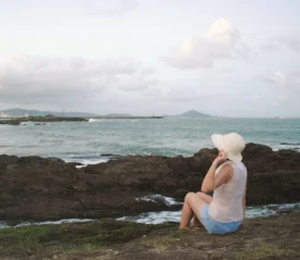Woman wearing a wide-brimmed hat seated on seaside rocks facing the distant water