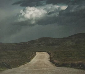 A dirt road winds through rolling hills toward dark storm clouds gathering overhead
