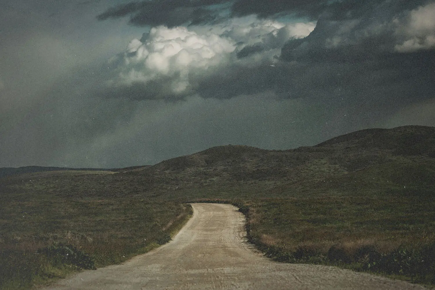 A dirt road winds through rolling hills toward dark storm clouds gathering overhead