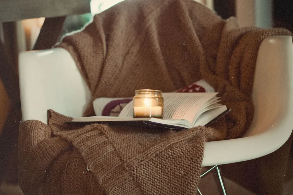 Cozy reading nook with a lit candle resting on an open book, draped with a chunky brown knit blanket over a white chair