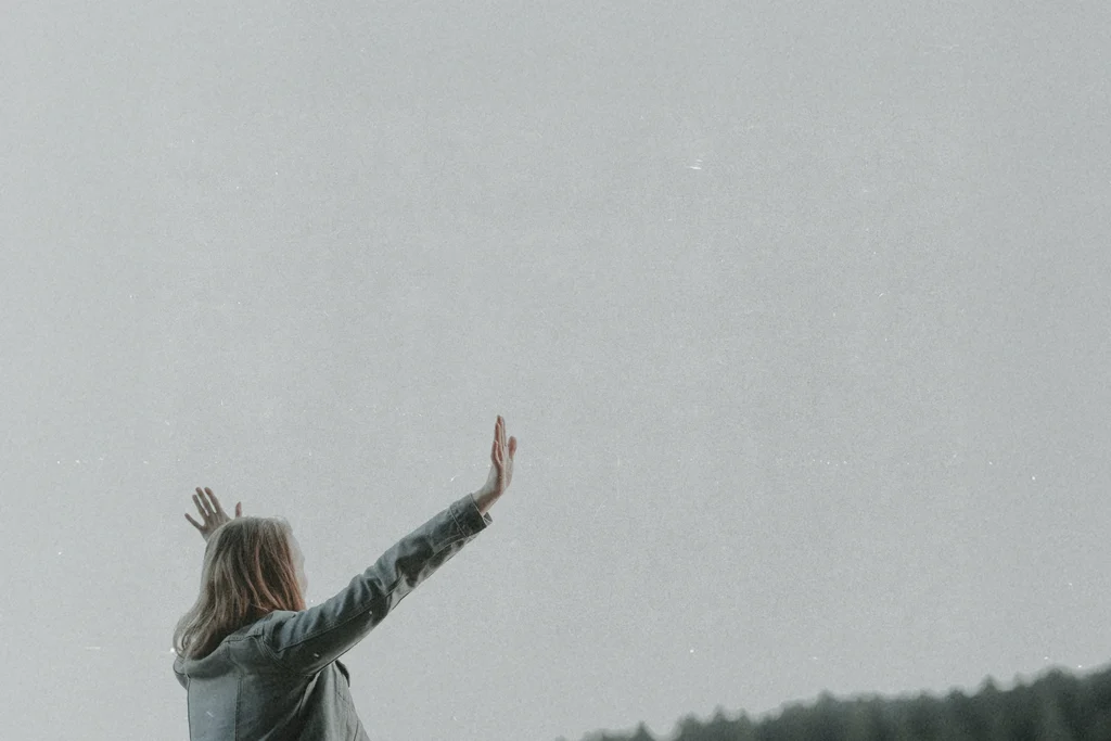 Person standing outdoors with arms raised toward the sky against a wide, soft gray backdrop and distant tree line