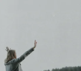 Person standing outdoors with arms raised toward the sky against a wide, soft gray backdrop and distant tree line