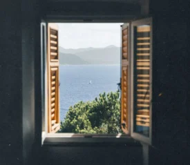 A window with wooden shutters opens to a scenic view of blue water, green treetops, and distant mountains