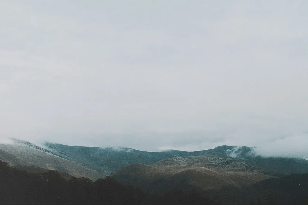 A series of low, rolling mountains are partially covered in mist beneath a gray, overcast sky
