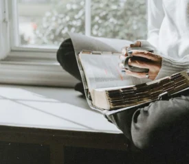 A person sits by a window holding a mug while reading an open Bible resting on their lap in soft natural light