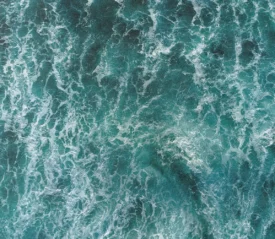 A top‑down view of churning ocean water shows swirling turquoise waves and white foam patterns