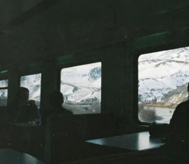 Passengers sit inside a dimly lit train car, looking out wide windows at snow‑covered mountains and a winding river