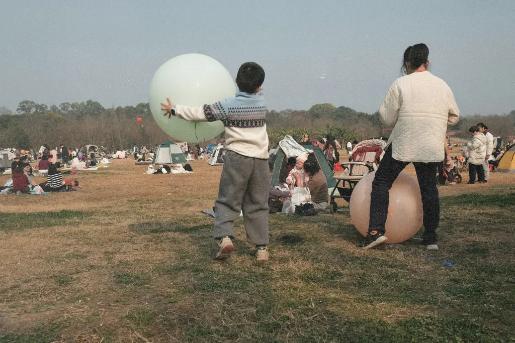 Children playing with large inflatable balls in an open grassy park filled with families, tents, and picnic groups on a clear day