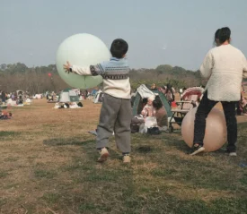 Children playing with large inflatable balls in an open grassy park filled with families, tents, and picnic groups on a clear day