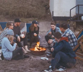 Group of friends sitting around a small beach bonfire, chatting and relaxing in deck chairs