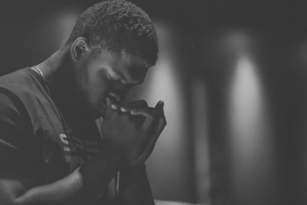 Person standing in a dimly lit room with hands raised near their face in a posture of reflection or prayer