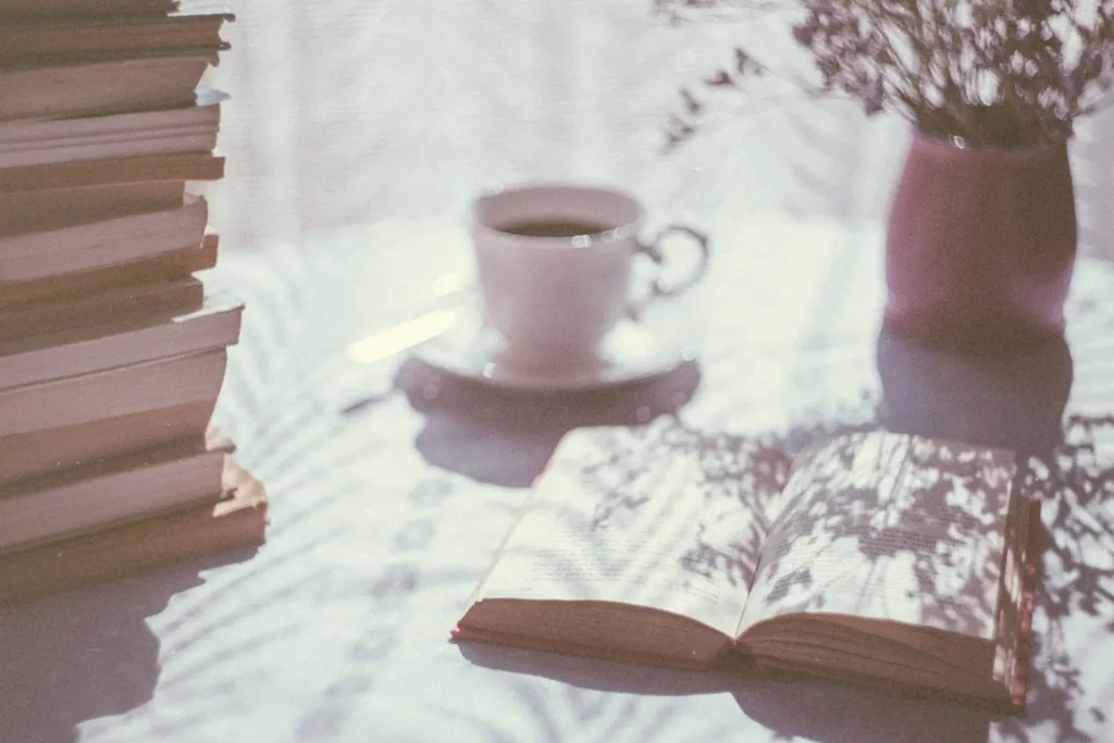 Open book lying on a sunlit table beside a stack of books, a cup of coffee, and a vase of dried flowers with soft shadows cast across the surface