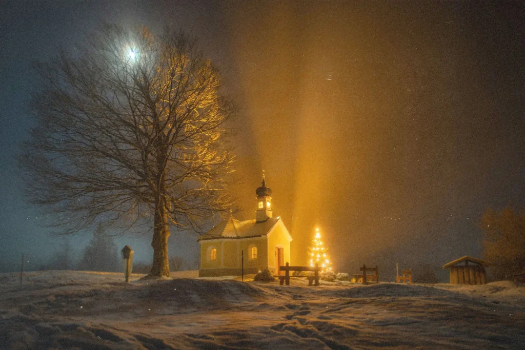 Snow‑covered church glowing with warm golden lights at night, with a lit Christmas tree and a large bare tree under a bright moon