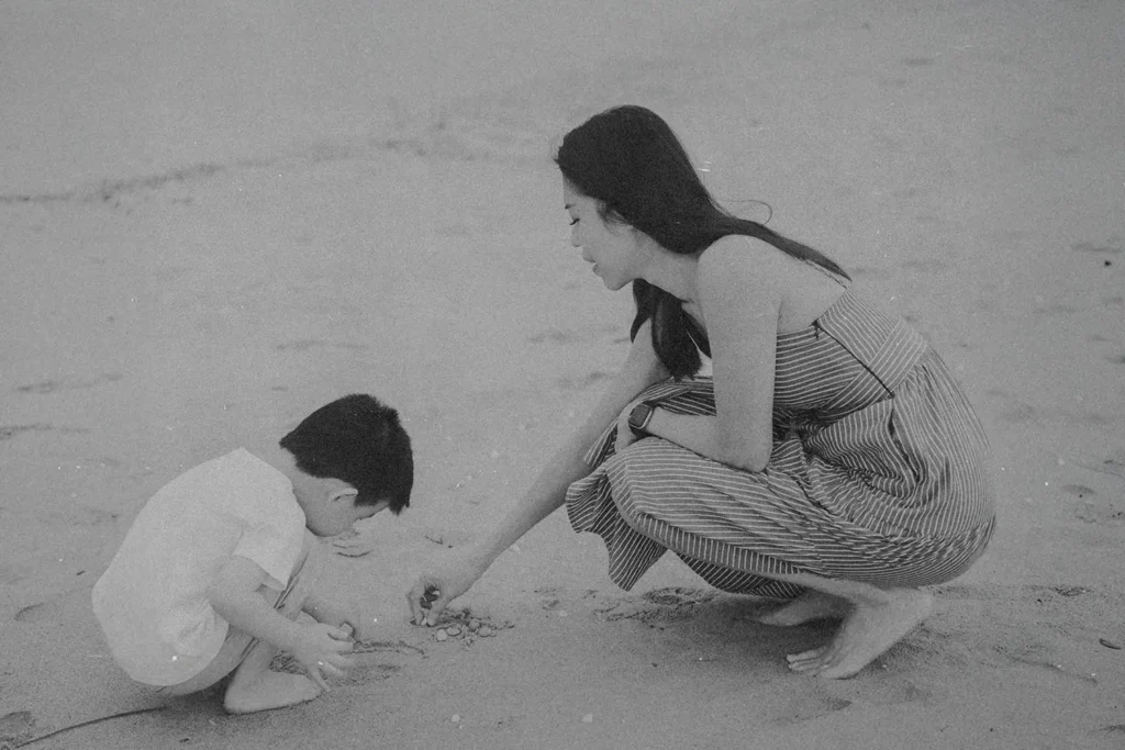 A woman kneels on the beach beside a child as they look closely at small objects in the sand
