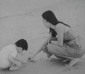 A woman kneels on the beach beside a child as they look closely at small objects in the sand