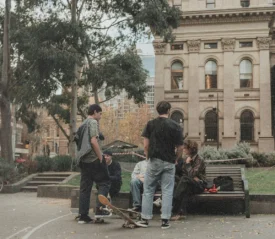 A group of people gather in an outdoor plaza near a bench, with skateboards nearby and historic buildings in the background