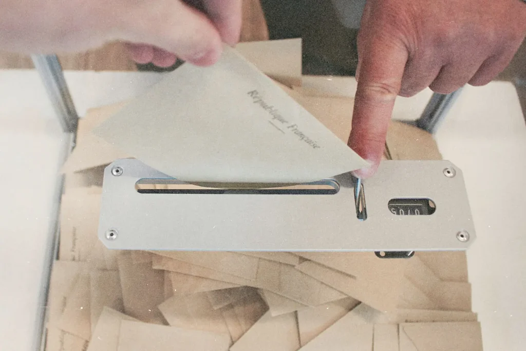 A hand places an envelope into a metal ballot box slot, with many sealed envelopes already collected inside