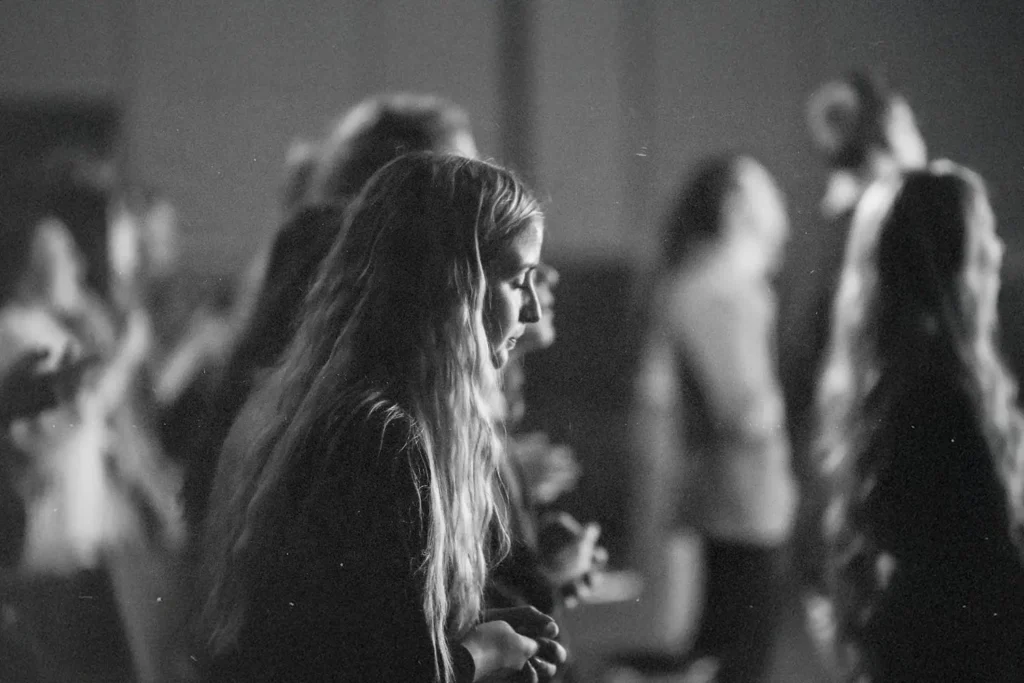 People standing in a dimly lit room during what appears to be a worship or group gathering, with individuals facing forward in soft focus