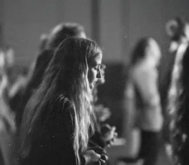 People standing in a dimly lit room during what appears to be a worship or group gathering, with individuals facing forward in soft focus