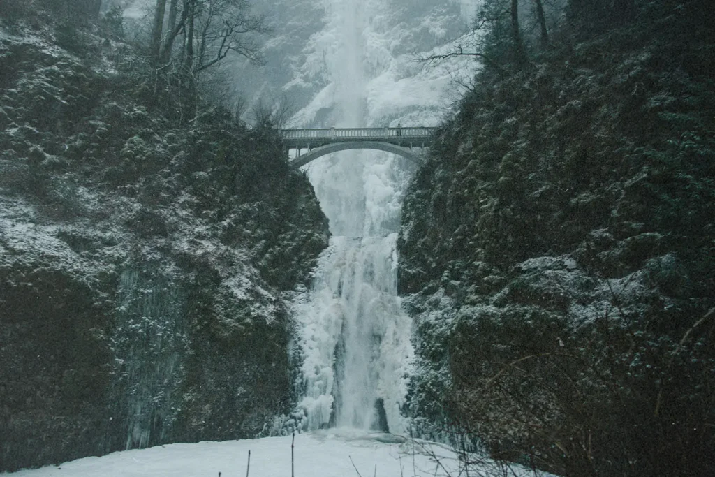Icy waterfall framed by rocky cliffs and a stone bridge overhead on a snowy day