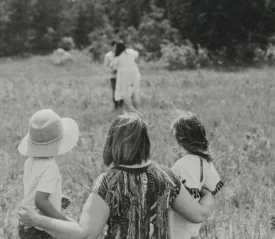 A woman stands in a grassy field with two young children at her sides, all facing toward another pair of people walking ahead across the open meadow, with trees lining the distance