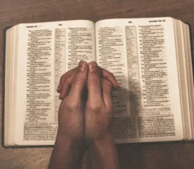 Hands folded in prayer over an open Bible on a wooden surface