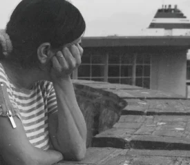 A person wearing a striped shirt leans on a brick wall, looking out toward a large docked ship in the distance