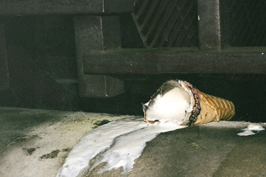 A dropped ice cream cone lies on the ground, with melted ice cream spreading across the surface near a dark metal structure