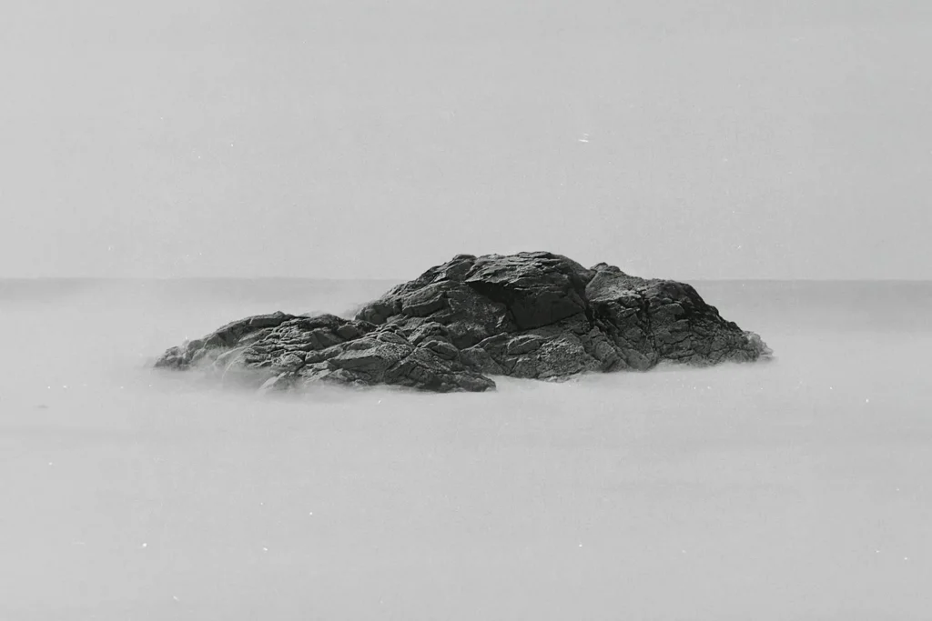 Large dark rock formation emerging from smooth, mist-like water under a pale, overcast sky