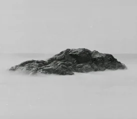 Large dark rock formation emerging from smooth, mist-like water under a pale, overcast sky