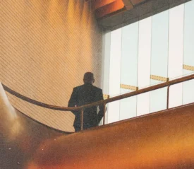 Figure leaning on a railing inside a contemporary structure with warm lighting and large windows