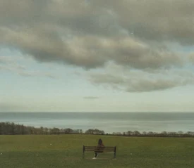 Person sitting on a bench in the middle of grassy field overlooking ocean view