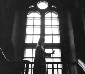 Silhouetted person standing indoors and touching a tall, bright window with arched panes, with light streaming in behind them