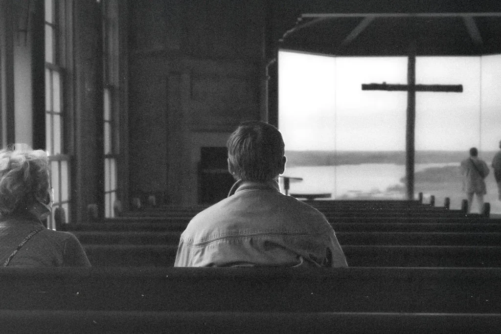 Two people sitting in church pews facing a large window with a cross silhouetted against a distant landscape, while others stand near the front