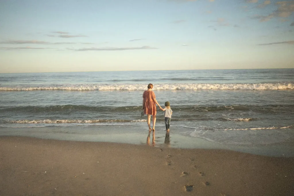 Adult and child standing side by side on the beach, looking out at the calm ocean under a pastel sky