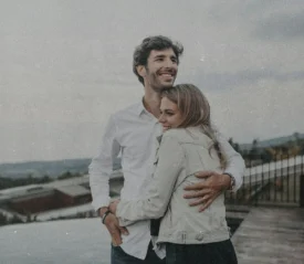 Two people stand together outdoors on a rooftop or terrace, with one person’s arm wrapped around the other as they look toward the landscape