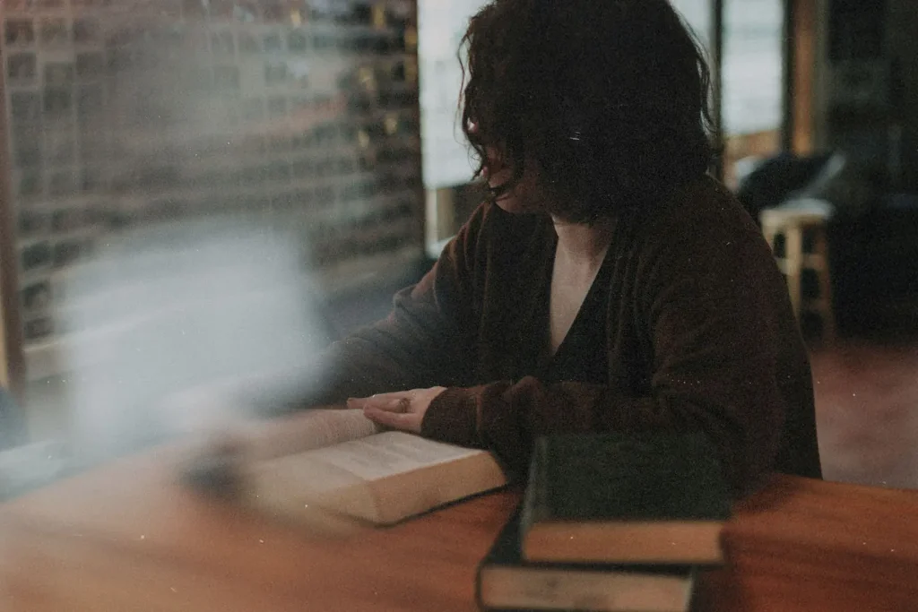 Person sitting at a wooden table reading an open book, with additional books stacked nearby in a softly lit room