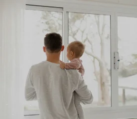 Adult holding a baby while standing by a large window, both looking outside at a softly lit, overcast landscape