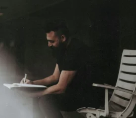 A person sits on a white outdoor chair, writing in a notebook, surrounded by deep shadows and soft light